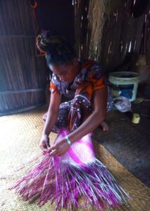 A woman is knelt on the floor and is weaving a basket with Mahampy grass. The grass is died bright pink and purple.