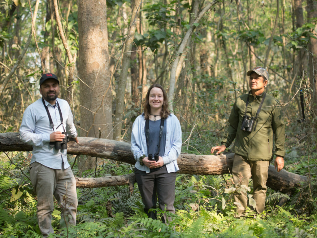 Three people stand in the middle of a dense forest, with a fallen tree trunk laying behind them. The people are holding binoculars and are smiling at the camera. 