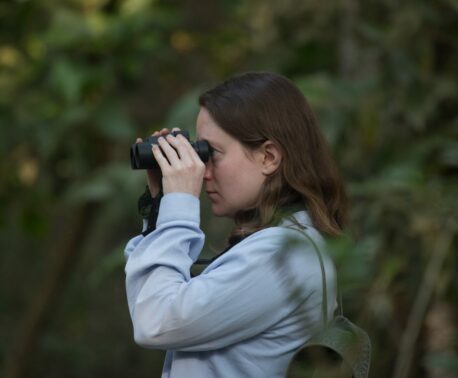 The headshot of a woman who stands in a dense forest looking through a pair of binoculars at something out of the shot.