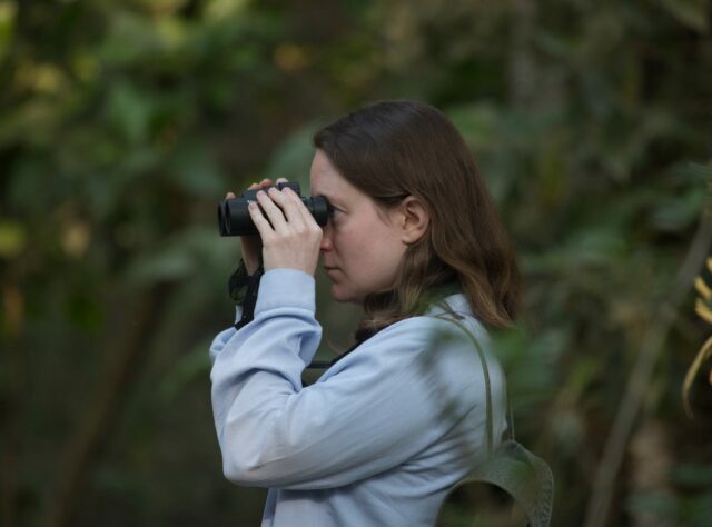 The headshot of a woman who stands in a dense forest looking through a pair of binoculars at something out of the shot.