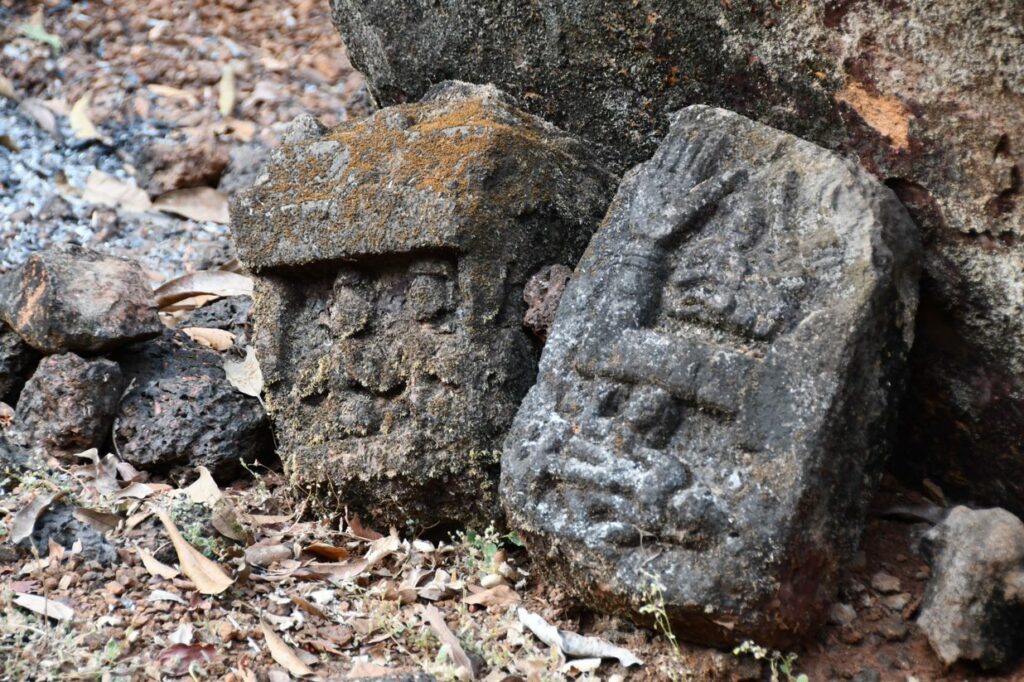 Two stones are leant against a larger rock with leaf litter beneath them. The stones are carved into, one with what appears to be a face and the other with shapes and markings. They are worn and appear very aged.