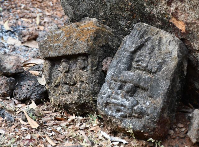 Two stones are leant against a larger rock with leaf litter beneath them. The stones are carved into, one with what appears to be a face and the other with shapes and markings. They are worn and appear very aged.