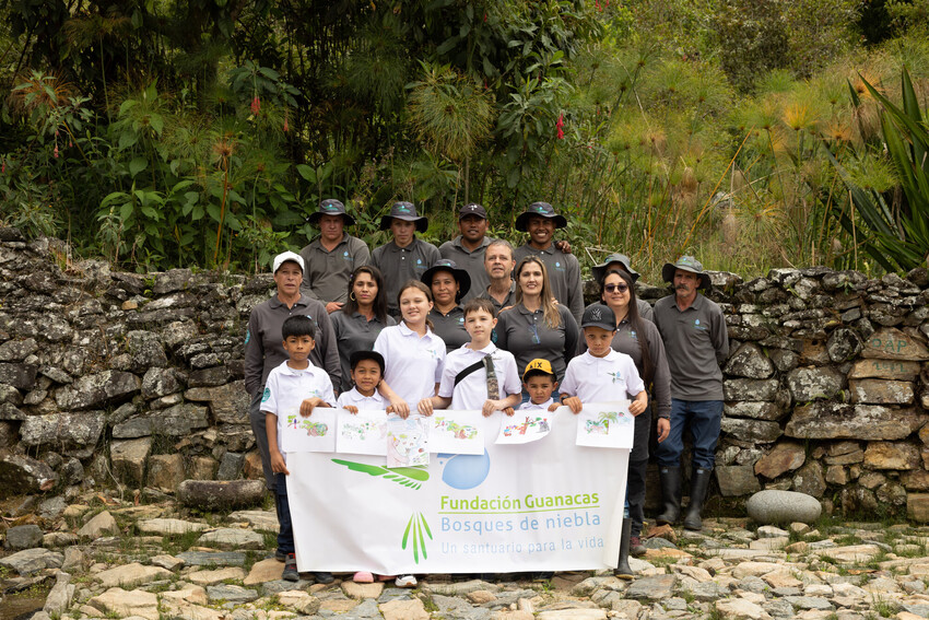A group of people stand together in front of an old stone wall with a forest in the background. The people are staff of the nature conservation charity Guanacas Foundation in Colombia and include forest rangers and their family members. 