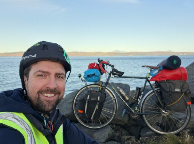 A person sits with a reflective tabard and waterproof coat by the sea on some rocks. A bicycle is in the background propped up against the rocks. The bicycle is loaded with panier bags and other items.