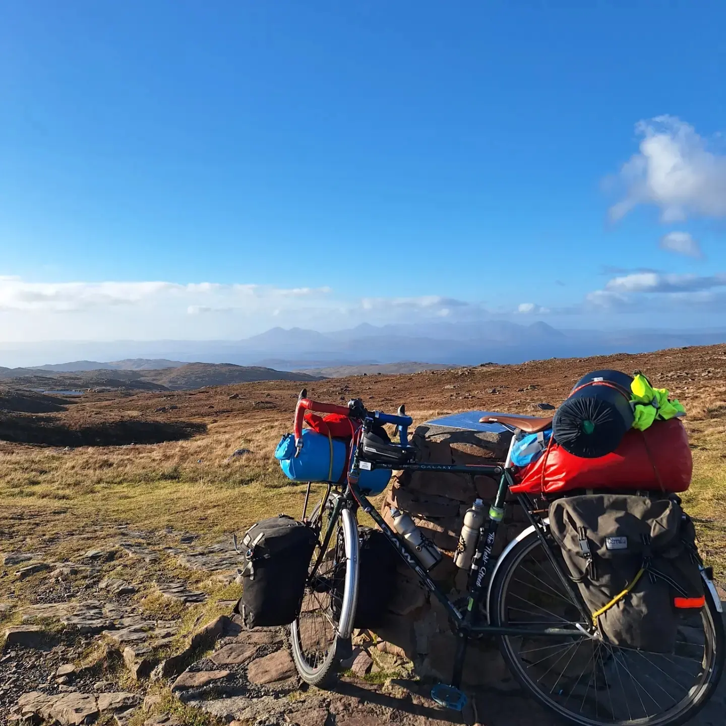 A bicycle full of cargo is propped up against a rock, with an open expanse of grassland behind and a clear blue sky.