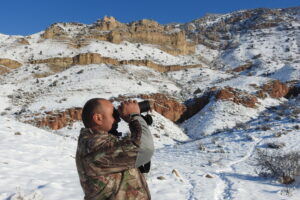 A ranger standing in front of hills and snow, using binoculars.