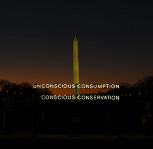 A neon sign with the words 'Unconscious Conservation; concious consumption' is stood in the foreground of a very tall thin obelisk which is the Washington D.C. National Mall and Monument. The sky is dark with a red hue at the horizon.