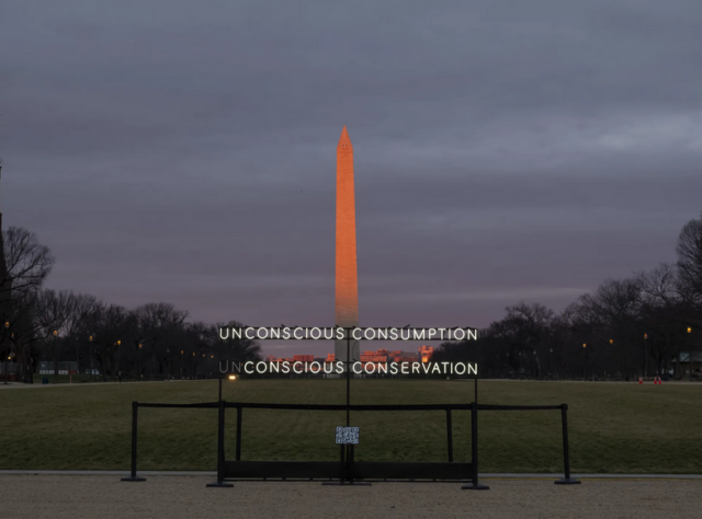A neon sign with the words 'Unconscious Conservation; concious consumption' is stood in the foreground of a very tall thin obelisk which is the Washington D.C. National Mall and Monument. The sky is dark with a red hue at the horizon.