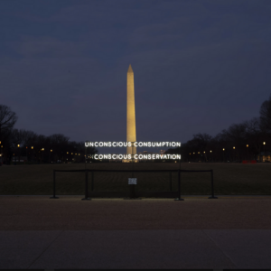 A neon sign with the words 'Unconscious Conservation; concious consumption' is stood in the foreground of a very tall thin obelisk which is the Washington D.C. National Mall and Monument.