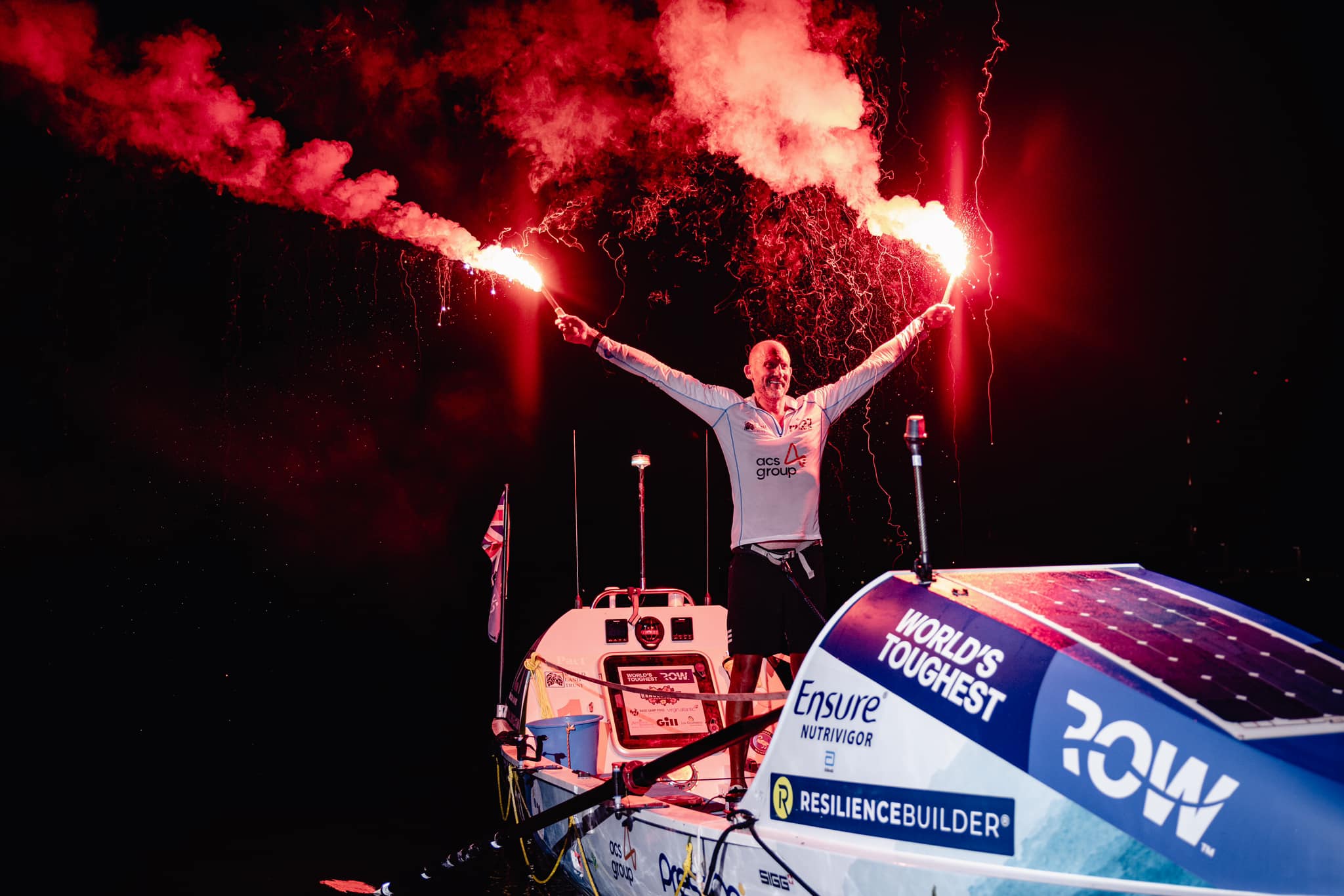 A man stands on a rowing boat in the darkness, lighting up the air with two red flares which he holds high in the air with his hands. The flares give off red tinted smoke.