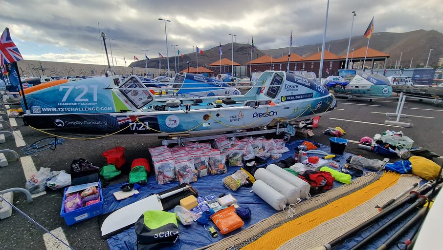 A rowing boat on land sits with an array of food, supplies, clothing and boat gear layed out on the floor infront of it.