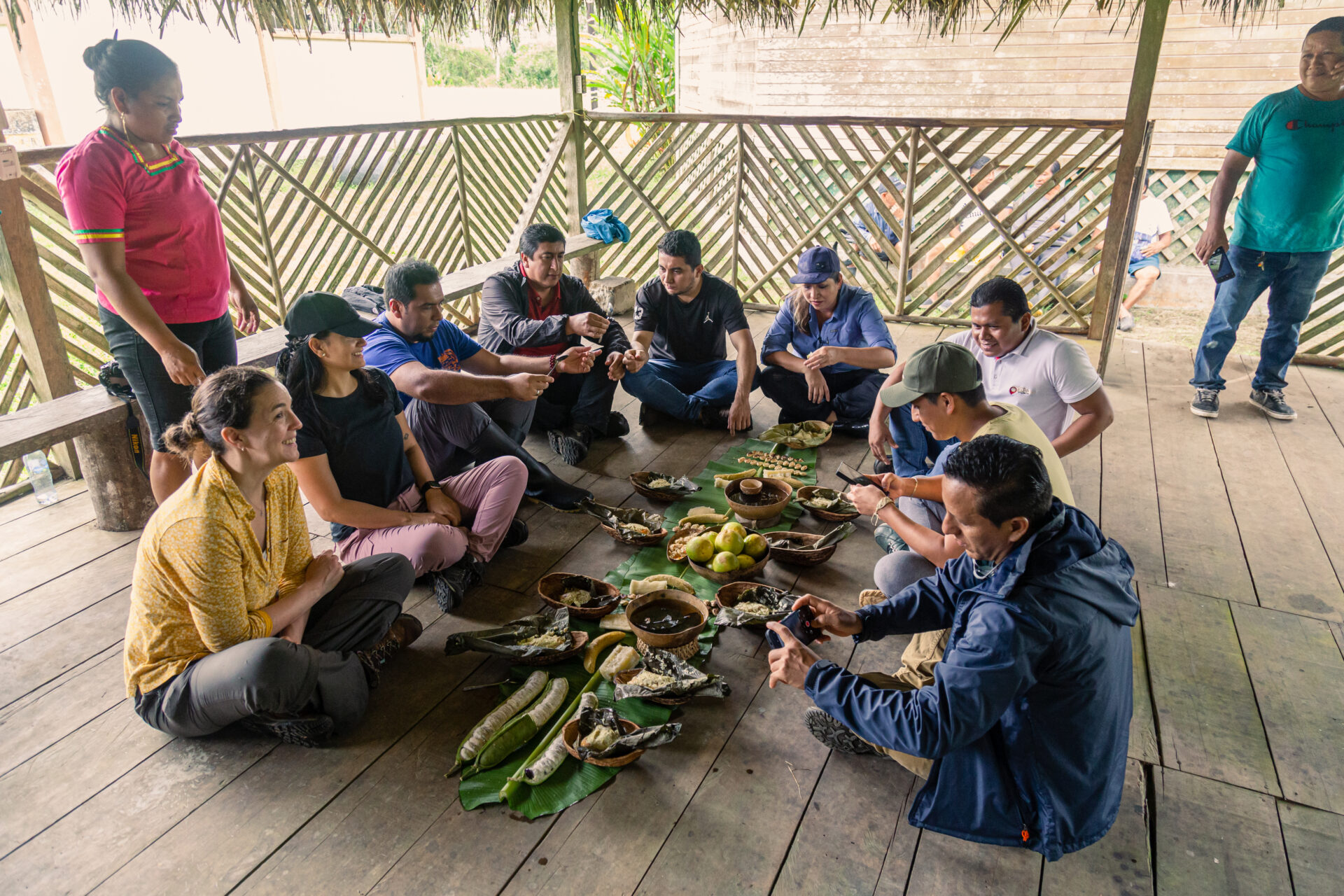 A group of people sitting on the floor of a wooden building enjoying a spread of traditional food from the Ecuadorian Amazon. 