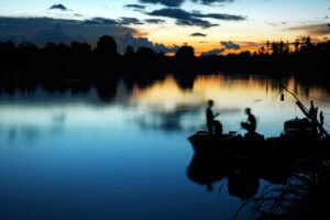 The Kinabatangan River at sunset with golden orange light in the distance mixing with the dusky blue of the water. In the foreground there are two men on a fishing boat.