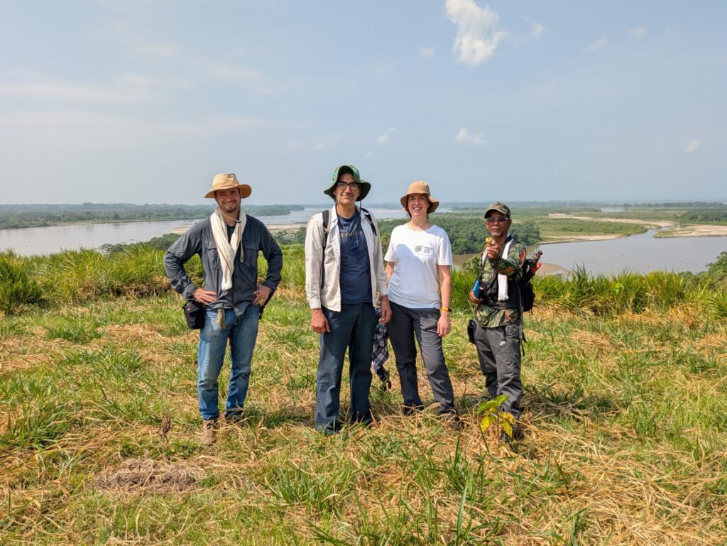 Four people stand together in a grassy lakeside landscape on a sunny day