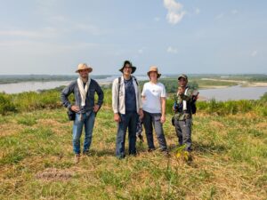 Four people stand together in a grassy lakeside landscape on a sunny day