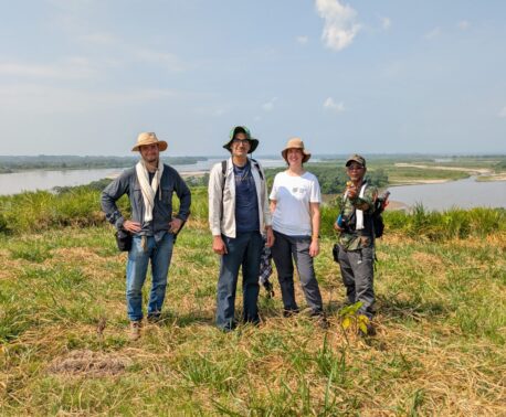 Four people stand together in a grassy lakeside landscape on a sunny day